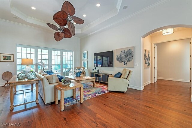 living room featuring a raised ceiling, wood finished floors, arched walkways, crown molding, and baseboards