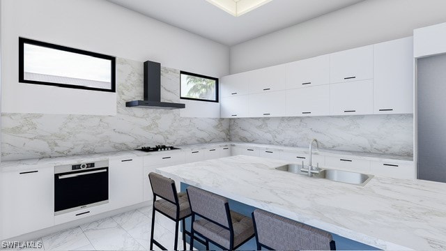 kitchen with wall chimney range hood, oven, white cabinets, light stone counters, and sink