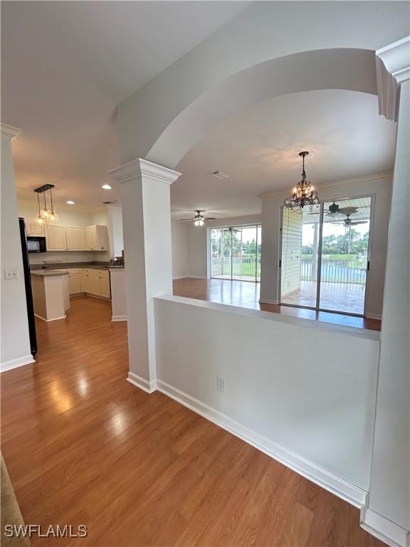 unfurnished room with arched walkways, ceiling fan with notable chandelier, wood finished floors, baseboards, and decorative columns