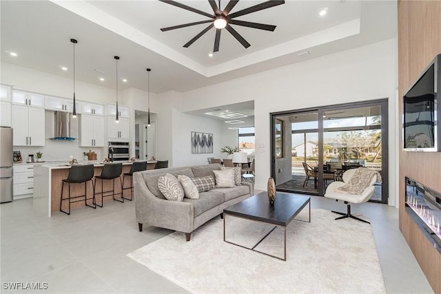 living area featuring recessed lighting, a glass covered fireplace, a towering ceiling, a ceiling fan, and a raised ceiling