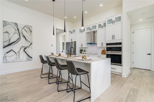 kitchen with white cabinetry, hanging light fixtures, an island with sink, stainless steel appliances, and wall chimney range hood