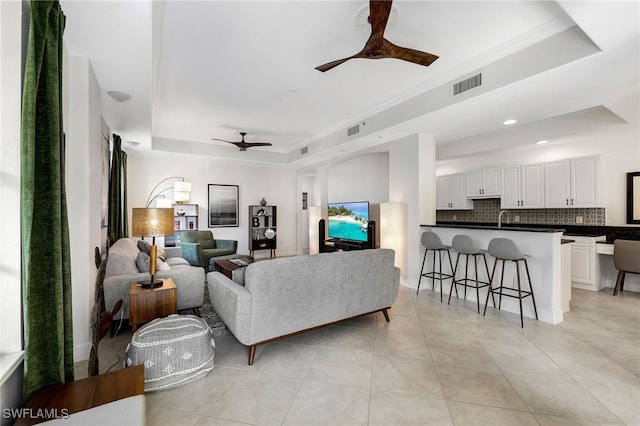 tiled living room with ceiling fan, crown molding, sink, and a tray ceiling