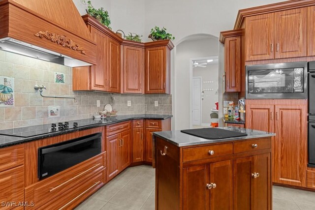 kitchen featuring dark stone countertops, backsplash, light tile patterned floors, black appliances, and custom range hood