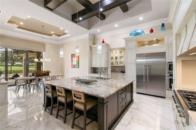 kitchen featuring light stone countertops, backsplash, built in appliances, a large island with sink, and a breakfast bar