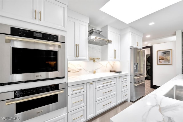 kitchen with appliances with stainless steel finishes, stacked washer and clothes dryer, white cabinets, and wall chimney range hood