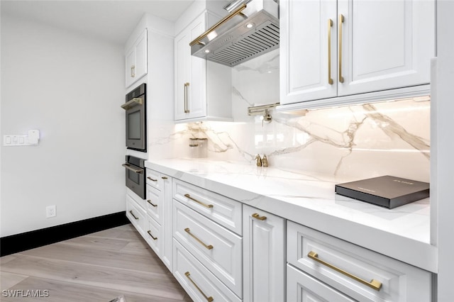 kitchen with wall chimney range hood, white cabinetry, light stone counters, and backsplash