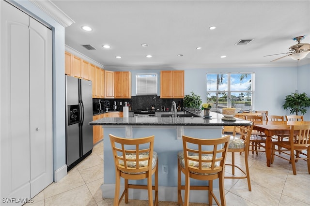 kitchen with backsplash, a center island with sink, stainless steel fridge with ice dispenser, ceiling fan, and dark stone countertops