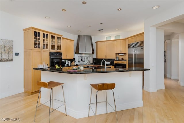 kitchen with built in fridge, glass insert cabinets, black oven, a large island with sink, and wall chimney exhaust hood