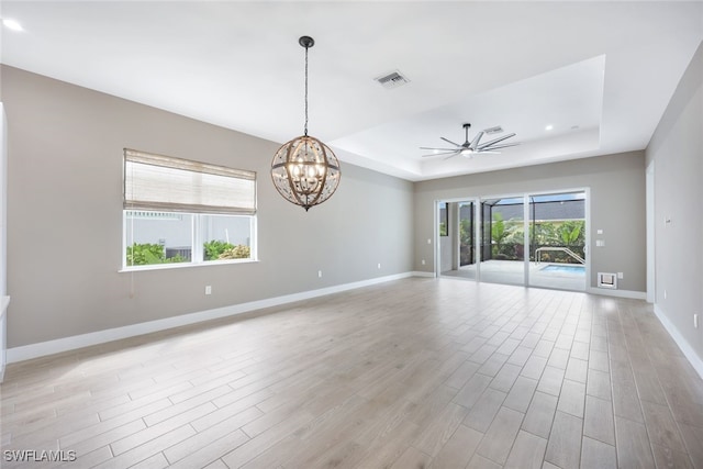 empty room featuring a tray ceiling, plenty of natural light, ceiling fan with notable chandelier, and light hardwood / wood-style flooring