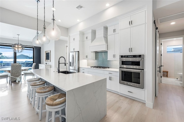 kitchen featuring decorative light fixtures, appliances with stainless steel finishes, premium range hood, an island with sink, and light wood-type flooring