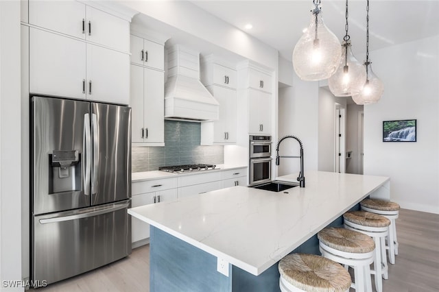 kitchen featuring decorative light fixtures, custom range hood, appliances with stainless steel finishes, an island with sink, and light wood-type flooring