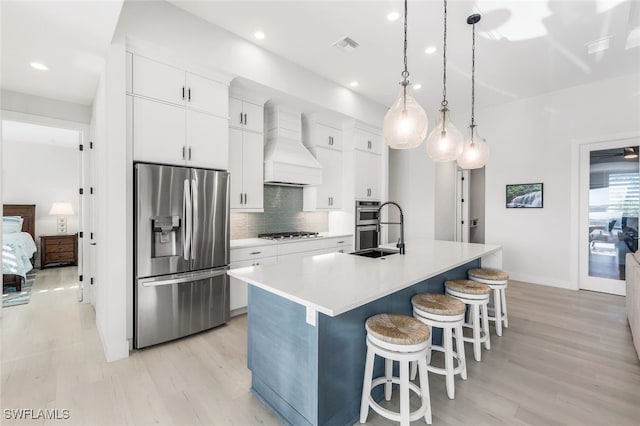 kitchen featuring stainless steel appliances, sink, custom range hood, a center island with sink, and white cabinets