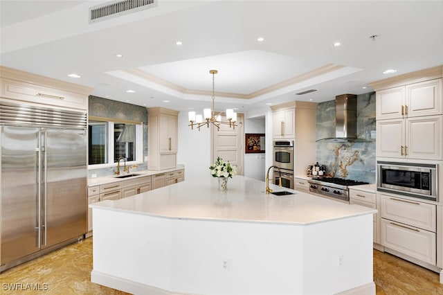 kitchen featuring visible vents, a sink, built in appliances, a raised ceiling, and wall chimney range hood