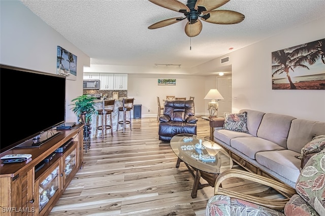 living room with a textured ceiling, light wood-style flooring, visible vents, baseboards, and a ceiling fan