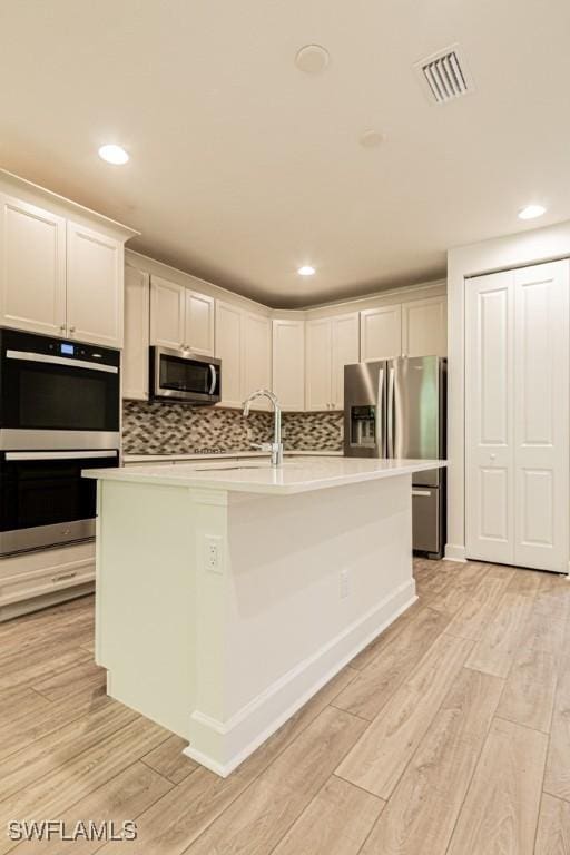 kitchen featuring appliances with stainless steel finishes, backsplash, a center island with sink, light hardwood / wood-style floors, and white cabinetry