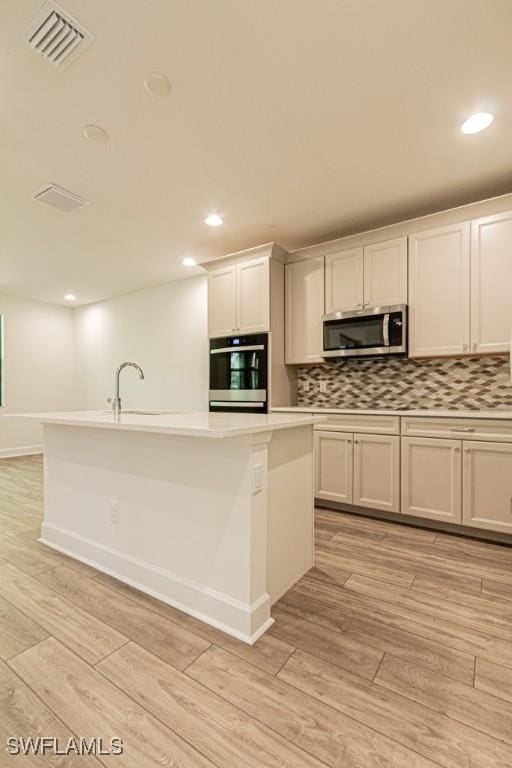 kitchen featuring a center island with sink, light wood-type flooring, appliances with stainless steel finishes, tasteful backsplash, and white cabinetry