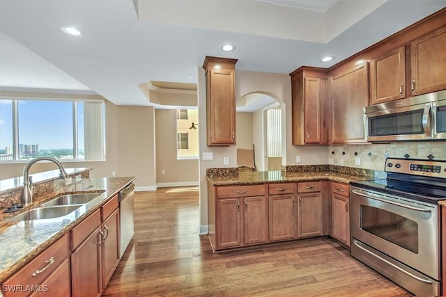 kitchen with a sink, a tray ceiling, dark stone counters, stainless steel appliances, and light wood-style flooring