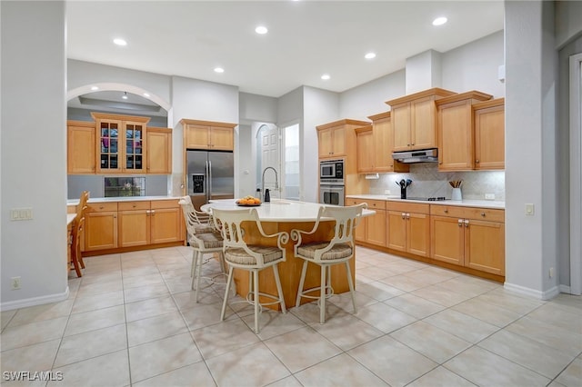 kitchen featuring a breakfast bar area, a center island with sink, light tile patterned floors, backsplash, and appliances with stainless steel finishes