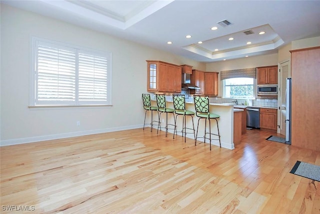 kitchen featuring a tray ceiling, brown cabinets, stainless steel appliances, wall chimney range hood, and a peninsula