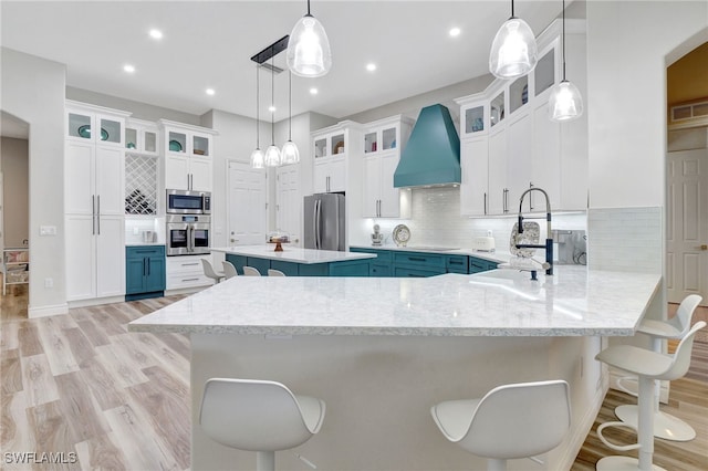 kitchen with light wood-type flooring, tasteful backsplash, decorative light fixtures, appliances with stainless steel finishes, and custom range hood