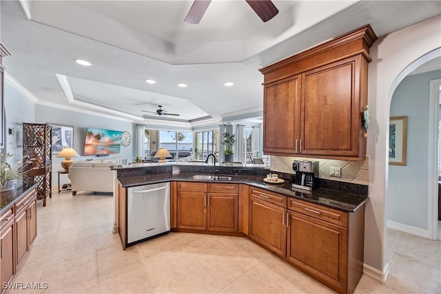 kitchen featuring dark stone counters, a raised ceiling, open floor plan, stainless steel dishwasher, and a sink