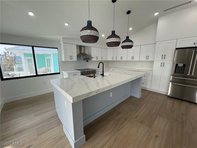 kitchen featuring white cabinets, wall chimney range hood, light stone countertops, an island with sink, and appliances with stainless steel finishes