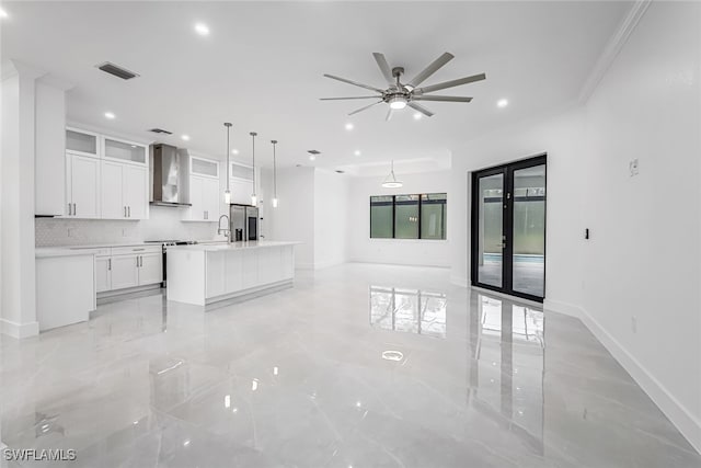 kitchen featuring ceiling fan, pendant lighting, wall chimney exhaust hood, a kitchen island with sink, and white cabinets