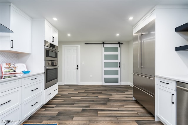 kitchen with white cabinets, a barn door, stainless steel appliances, wall chimney range hood, and dark hardwood / wood-style floors