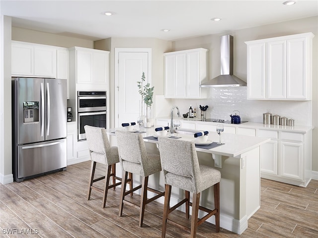 kitchen featuring an island with sink, stainless steel appliances, wall chimney range hood, and white cabinetry