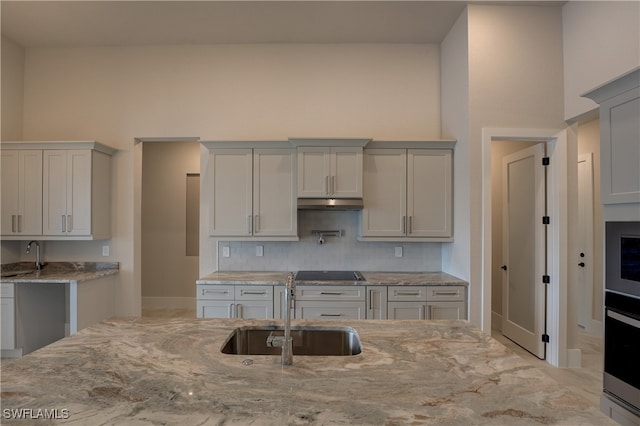 kitchen with black electric stovetop, sink, backsplash, stainless steel oven, and light stone counters