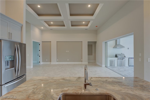 kitchen with a towering ceiling, sink, wall chimney range hood, light stone countertops, and stainless steel fridge with ice dispenser