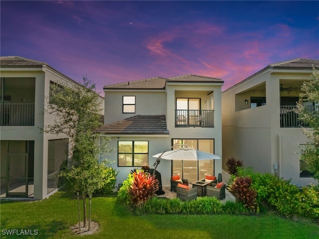 back house at dusk featuring a balcony, an outdoor hangout area, a lawn, and a patio area