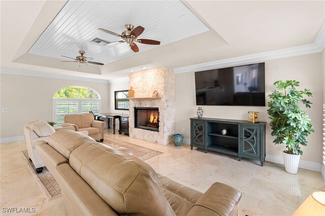 living room with ceiling fan, a raised ceiling, a stone fireplace, crown molding, and light tile patterned flooring