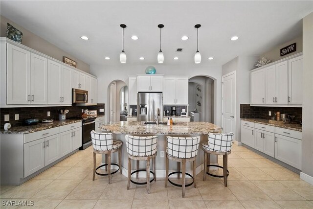 kitchen with a kitchen island with sink, white cabinetry, stainless steel appliances, decorative light fixtures, and dark stone countertops