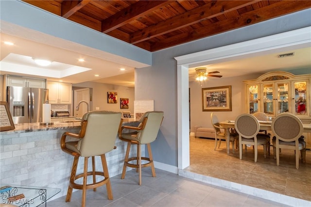 kitchen featuring sink, a breakfast bar, stainless steel refrigerator with ice dispenser, light stone counters, and wooden ceiling