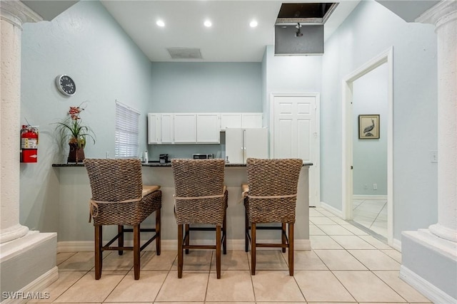 interior space with dark countertops, freestanding refrigerator, ornate columns, a kitchen bar, and white cabinetry