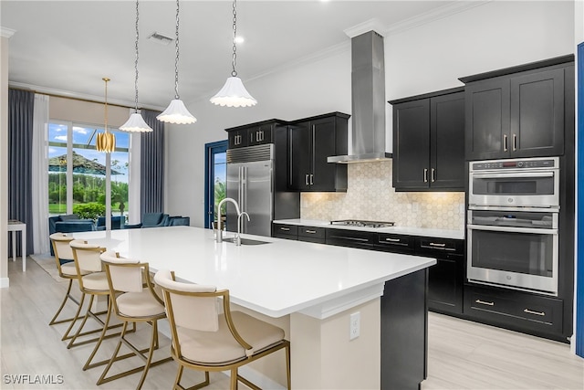 kitchen featuring backsplash, appliances with stainless steel finishes, an island with sink, hanging light fixtures, and wall chimney range hood