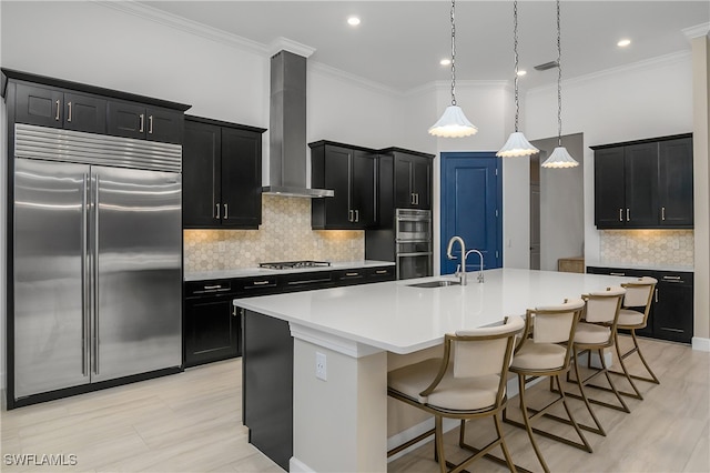 kitchen featuring wall chimney exhaust hood, a kitchen bar, decorative light fixtures, a center island with sink, and stainless steel appliances