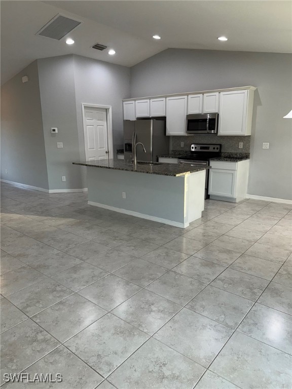 kitchen featuring a center island with sink, vaulted ceiling, dark stone countertops, white cabinetry, and stainless steel appliances