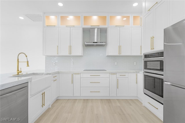 kitchen with light wood-type flooring, light stone counters, stainless steel appliances, wall chimney range hood, and white cabinets