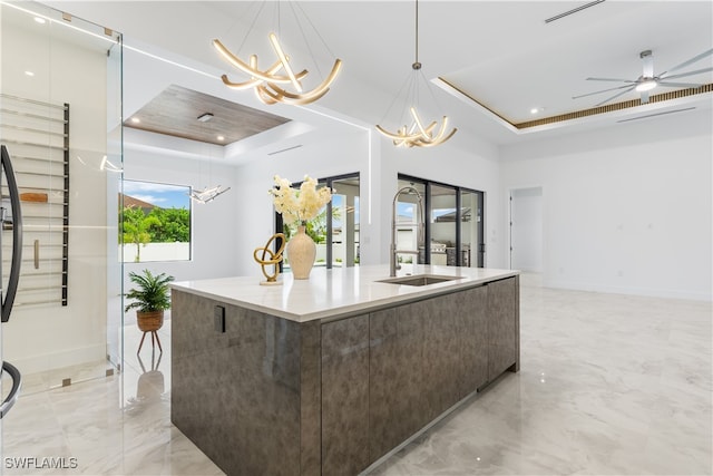 kitchen featuring dark brown cabinetry, a raised ceiling, an island with sink, decorative light fixtures, and ceiling fan with notable chandelier