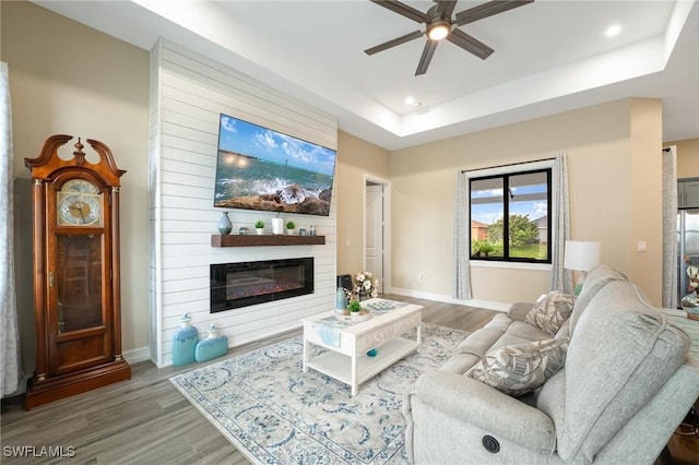 living room featuring wood-type flooring, a large fireplace, a tray ceiling, and ceiling fan