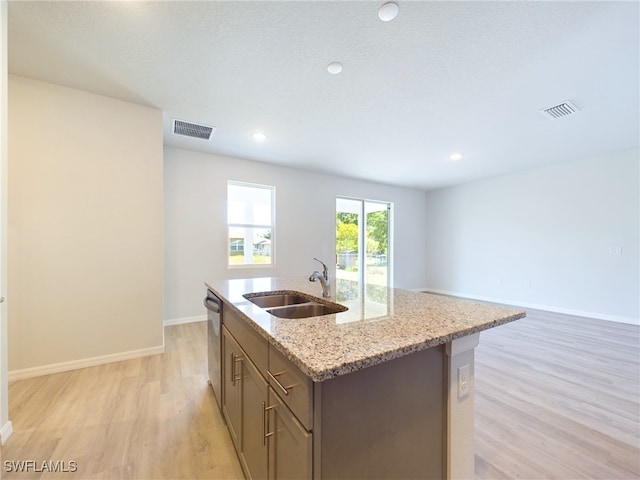 kitchen with light wood-type flooring, stainless steel dishwasher, light stone countertops, sink, and a center island with sink