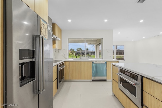 kitchen featuring light tile patterned flooring, backsplash, appliances with stainless steel finishes, light stone countertops, and light brown cabinetry