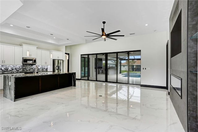 kitchen featuring white cabinetry, a raised ceiling, an island with sink, ceiling fan, and stainless steel appliances