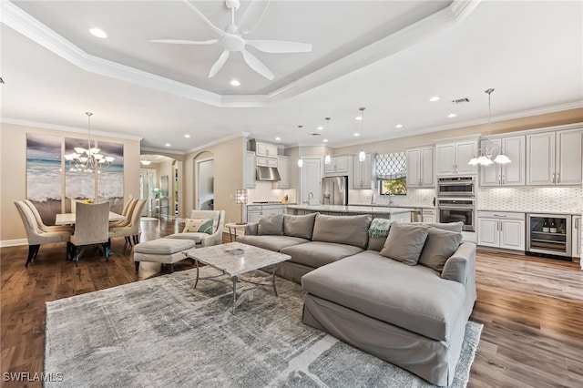 living room featuring wine cooler, ceiling fan with notable chandelier, a raised ceiling, and wood finished floors