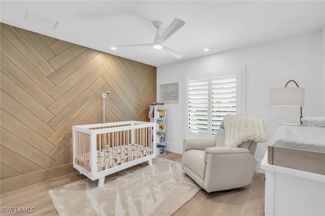 bedroom featuring a nursery area, ceiling fan, wooden walls, and wood finished floors