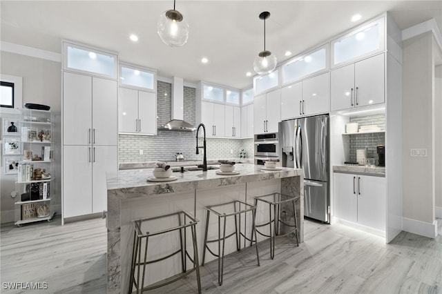 kitchen with white cabinetry, stainless steel appliances, wall chimney range hood, backsplash, and decorative light fixtures