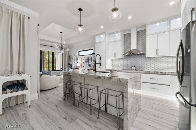 kitchen featuring backsplash, wall chimney exhaust hood, white cabinetry, stainless steel refrigerator, and an island with sink