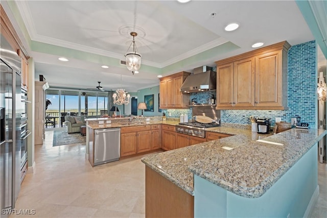 kitchen featuring appliances with stainless steel finishes, decorative light fixtures, a peninsula, a tray ceiling, and wall chimney range hood